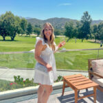 woman standing outside in white dress with drink in hand.
