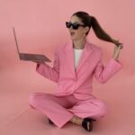 woman sitting in pink suit with sunglasses and a laptop pulling her hair.
